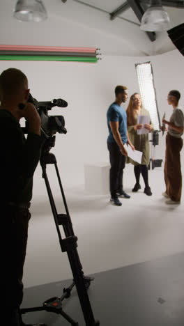 Vertical-Video-Of-Film-Camera-Operator-Shooting-Director-Talking-With-Male-And-Female-Actors-Holding-Scripts-Rehearsing-For-Shooting-Movie-Or-Video-In-Studio-2